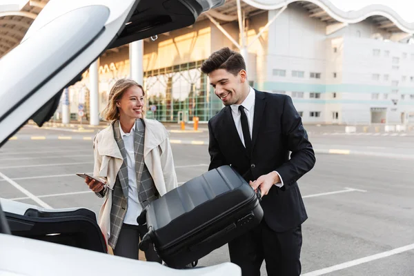 Image of young businesslike man and woman putting luggage in car — 图库照片