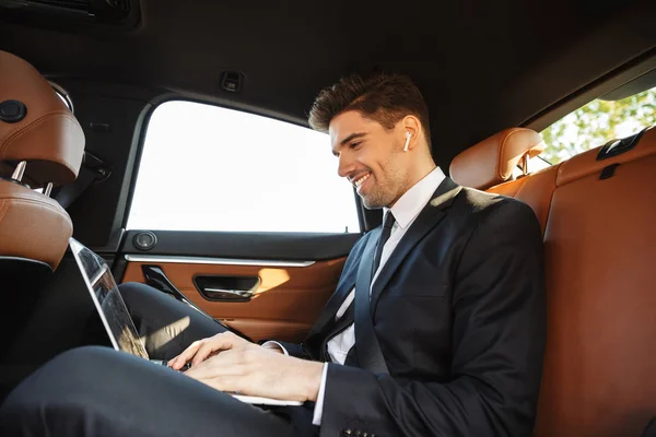 Foto de hombre de negocios sonriente usando auricular y portátil mientras va en coche —  Fotos de Stock