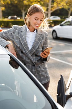 Photo of businesswoman using earpod and cellphone while leaning on car