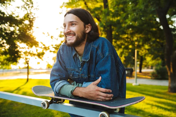 Positiv optimistisch lächelnder junger bärtiger Mann mit Skateboard steht im Naturpark. — Stockfoto