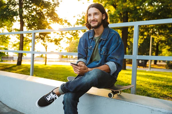 Happy smiling young bearded man with skateboard sit in nature park using mobile phone. — Stock Photo, Image