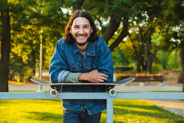 Glimlachende optimistische jonge man met baard en skateboard in het natuurpark. — Stockfoto