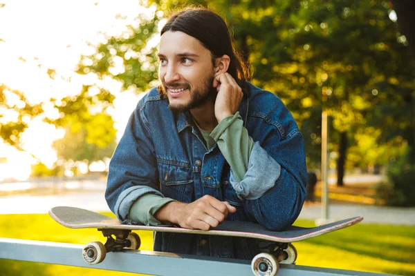 Usmívající se optimistický mladý vousatý muž se skateboardem stojící v přírodním parku. — Stock fotografie