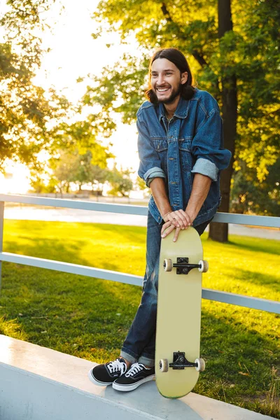 Glimlachende optimistische jonge man met baard en skateboard in het natuurpark. — Stockfoto