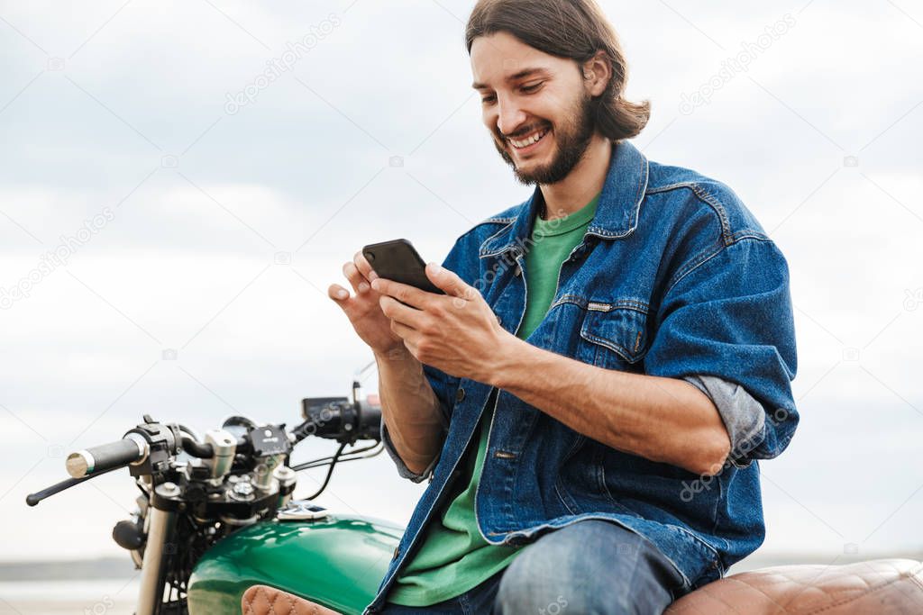 Pleased happy man biker on his bike outdoors on a beach using mobile phone chatting.