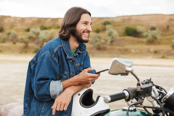 Piacevole felice uomo motociclista sulla sua moto all'aperto su una spiaggia utilizzando il telefono cellulare chat . — Foto Stock