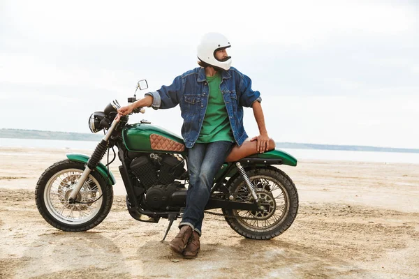 Homem motociclista em sua bicicleta ao ar livre em uma praia em um capacete . — Fotografia de Stock