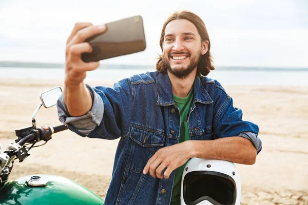 Bello sorridente compiaciuto uomo motociclista sulla sua moto all'aperto su una spiaggia farsi selfie dal telefono cellulare . — Foto Stock