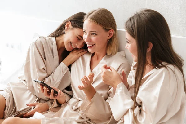 Three cheerful girls friends wearing dressing gowns sitting on bed — 스톡 사진