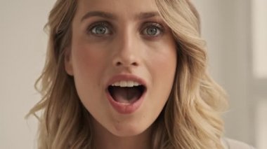 Sarışın, şaşırtıcı mutlu kadının yakın görüntüsü beyaz stüdyoda göz kırparken kameraya sessiz bir jest yapıyor.