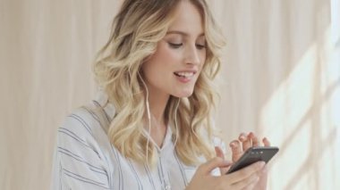 Neşeli, gülümseyen, güzel bir genç kadın, kumaş arka planında izole edilmiş telli kulaklıklarla müzik dinlerken akıllı telefon kullanıyor.