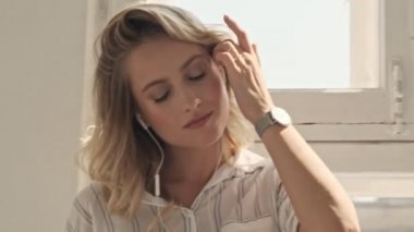 Güzel, gülümseyen bir kadın açık pencerelerin yanındaki beyaz ofiste akıllı telefonuyla sarı saçlarına dokunuyor.