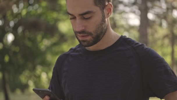 屋外の公園でスマートフォンを使用している間に何かを考え 周りを見回す高価なハンサムなスポーツマン — ストック動画