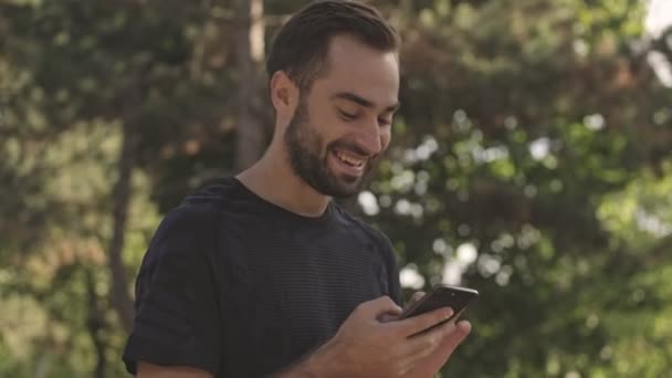 公園の屋外でスマートフォンを使用している間 陽気なハンサムなスポーツマンが歩き回って見て回る — ストック動画