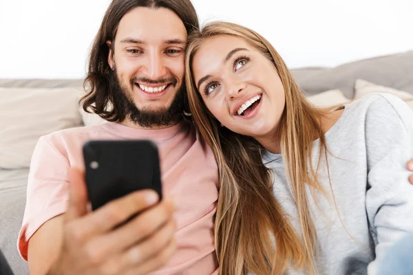 Loving couple on sofa indoors at home using mobile phone. — Stock Photo, Image