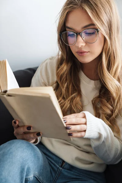 Foto de jovem mulher séria lendo livro enquanto sentado no sofá — Fotografia de Stock