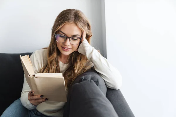 Foto van jonge tevreden vrouw die boek leest terwijl ze op de bank zit — Stockfoto