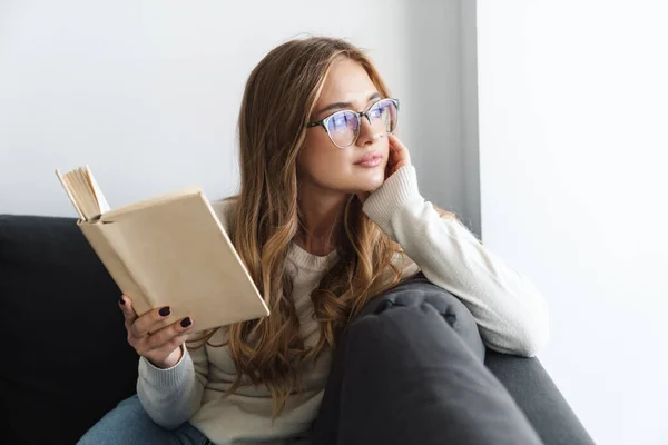 Fénykép fiatal komoly nő olvasás könyvet, miközben ül a kanapén — Stock Fotó