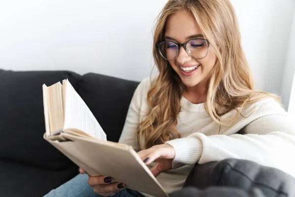 Φωτογραφία της νεαρής χαμογελαστής γυναίκας που διαβάζει το βιβλίο ενώ κάθεται στον καναπέ — Φωτογραφία Αρχείου