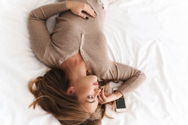 Genç ve güzel bir kadının akıllı telefonuyla yatakta yattığı fotoğraf.