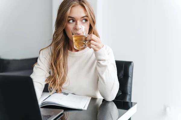 年轻的高加索女人喝茶和在笔记本电脑上打字的照片 — 图库照片