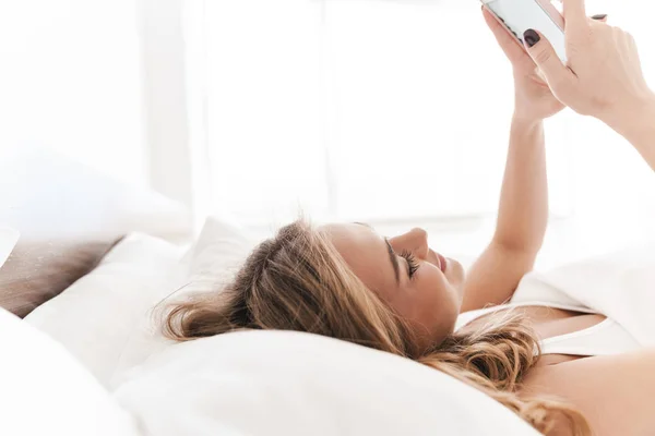 Foto de mujer caucásica alegre sonriendo y usando el teléfono celular mientras que miente —  Fotos de Stock