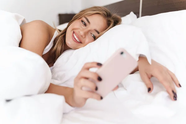 Foto de una mujer caucásica sonriente usando un teléfono celular mientras está acostada en la cama —  Fotos de Stock