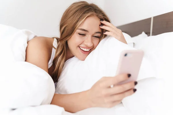 Foto de una mujer caucásica sonriente usando un teléfono celular mientras está acostada en la cama — Foto de Stock