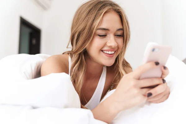 Foto de una mujer caucásica sonriente usando un teléfono celular mientras está acostada en la cama —  Fotos de Stock
