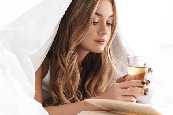 Foto van jonge gefocuste vrouw die boek leest en thee drinkt terwijl ze ligt — Stockfoto