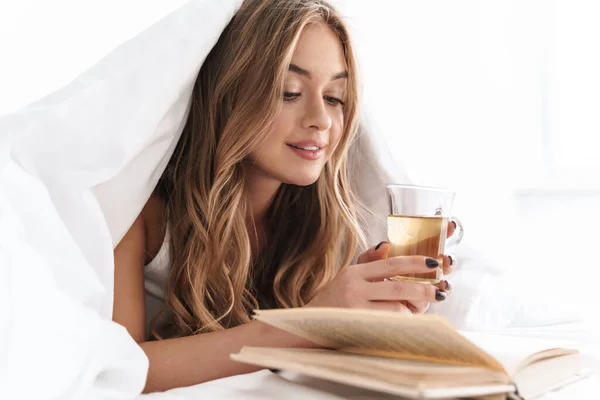 年轻女子躺在床上看书喝茶的照片 — 图库照片