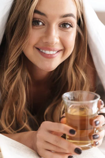 Foto de una joven mujer sonriente leyendo libro y tomando té mientras está acostada —  Fotos de Stock