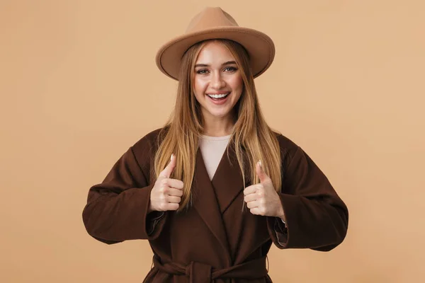 Obraz młodej dziewczyny w kapeluszu i płaszczu uśmiechnięty i pokazując thu — Zdjęcie stockowe
