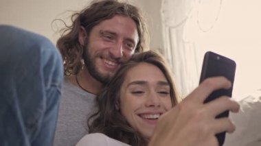 Evdeki yatağın yanında otururken akıllı telefon kullanan neşeli çiftin yakın görüntüsü.