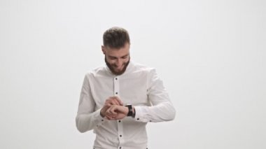 Beyaz tişörtlü, şık, genç, sakallı bir adam kameraya doğru yürürken beyaz duvar arkasında izole edilmiş akıllı saatini kullanıyor.