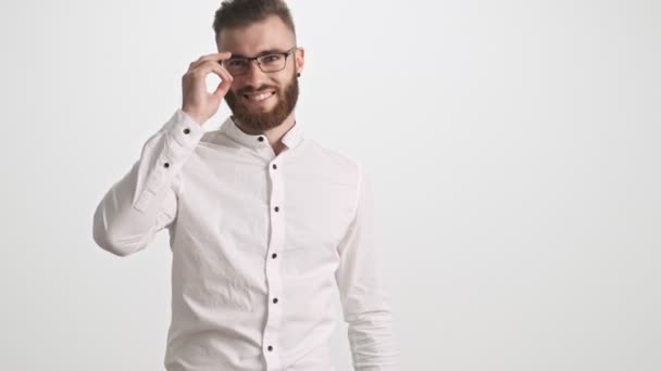 白いシャツを着た魅力的な若い髭の男が白い壁の背景に孤立した鼻の橋の上にメガネを置く — ストック動画