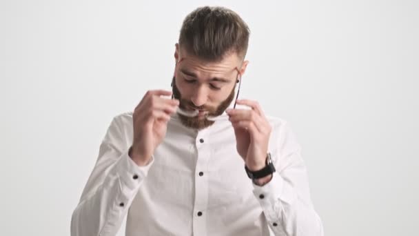 一个疲惫的留着胡子的年轻人摘下眼镜 触摸鼻梁 解开被白墙背景隔开的衬衫上的钮扣 — 图库视频影像