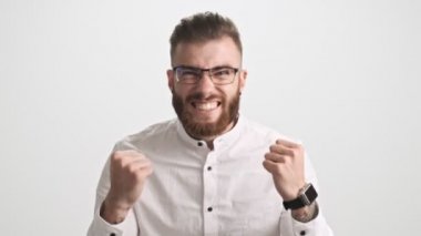 Beyaz tişörtlü, çekici, mutlu, sakallı bir adam beyaz duvar arkasına izole edilmiş bir jest yapıyor.
