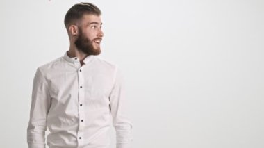 Beyaz tişörtlü, sakallı, şaşırmış genç bir adam büyük parmağını beyaz duvar arkasında izole edilmiş bir şekilde kenarı gösteriyor.