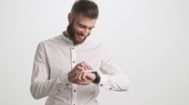 Gülümseyen genç sakallı bir adam beyaz duvar arkasında izole edilmiş akıllı saatini kullanıyor.