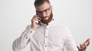Beyaz gömlek ve gözlük takan genç sakallı bir adam telefon görüşmesi sırasında beyaz duvar arkasında münakaşa ediyor.