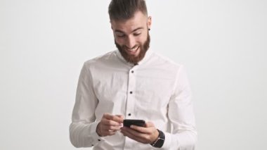 Beyaz gömlek giyen, gülümseyen, sakallı genç bir adam telefonunda ilginç bir şey olduğunu gösteriyor.
