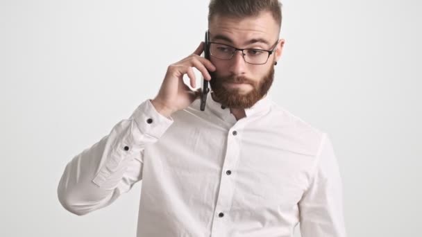 一个头戴白衬衫 戴着眼镜 严肃而沉默寡言的年轻大胡子男人 一边在白墙的背景上打了个电话 一边停止听电话 — 图库视频影像
