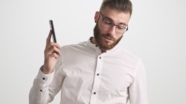 一个头戴白衬衫 戴着眼镜的大胡子的年轻人 在白墙的背景下完成了一个与外界隔绝的电话通话 — 图库视频影像