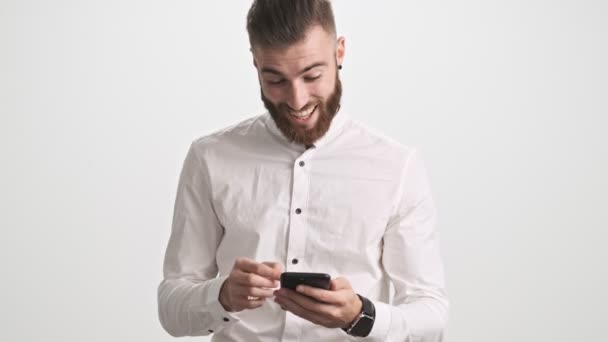 白いシャツを着た笑顔の若い髭の男は 白い壁の背景に隔離されたまま携帯電話に何か面白いものがあることを示しています — ストック動画