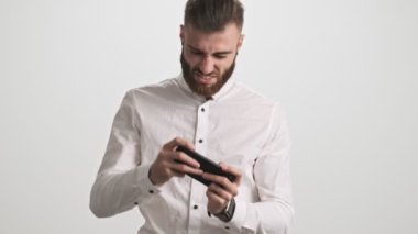 Beyaz tişörtlü neşeli, pozitif sakallı bir adam akıllı telefonuyla oyun oynuyor ve beyaz duvar arkasında tek başına kazanıyor.