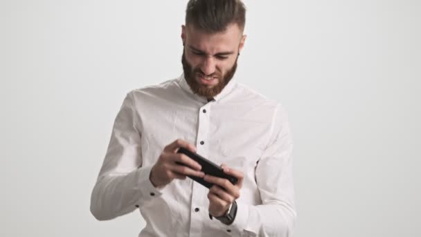 一位快乐的 积极的 留着白胡子的年轻人 穿着白衬衫 正在用他的智能手机玩游戏 然后在白墙的背景下赢得了一场比赛 — 图库视频影像