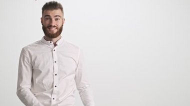Beyaz tişörtlü, gülümseyen genç sakallı bir adam iki eli ve ağzı açık bir şekilde beyaz duvar arkasına izole edilmiş bir kenarı işaret ediyor.