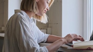A focused young woman is working on a laptop while sitting at the desk near the window