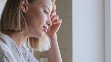 A pretty blonde young woman is using a laptop while sitting at the desk near the window